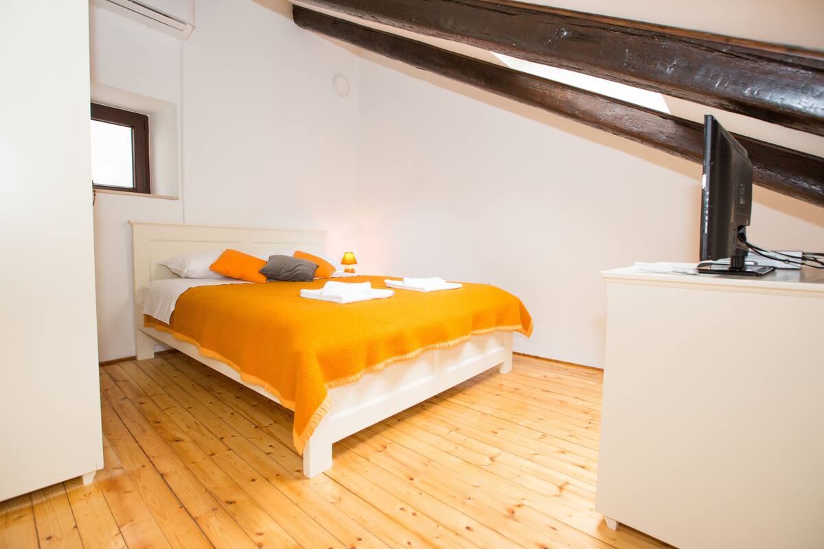 Un dormitorio con piso de madera, una cama con frazada naranja, almohadas blancas, una mesita de noche y un televisor sobre un mueble.
