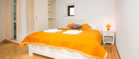 Un dormitorio con cama blanca, ropa de cama naranja, una mesita de noche con lámpara y un armario.