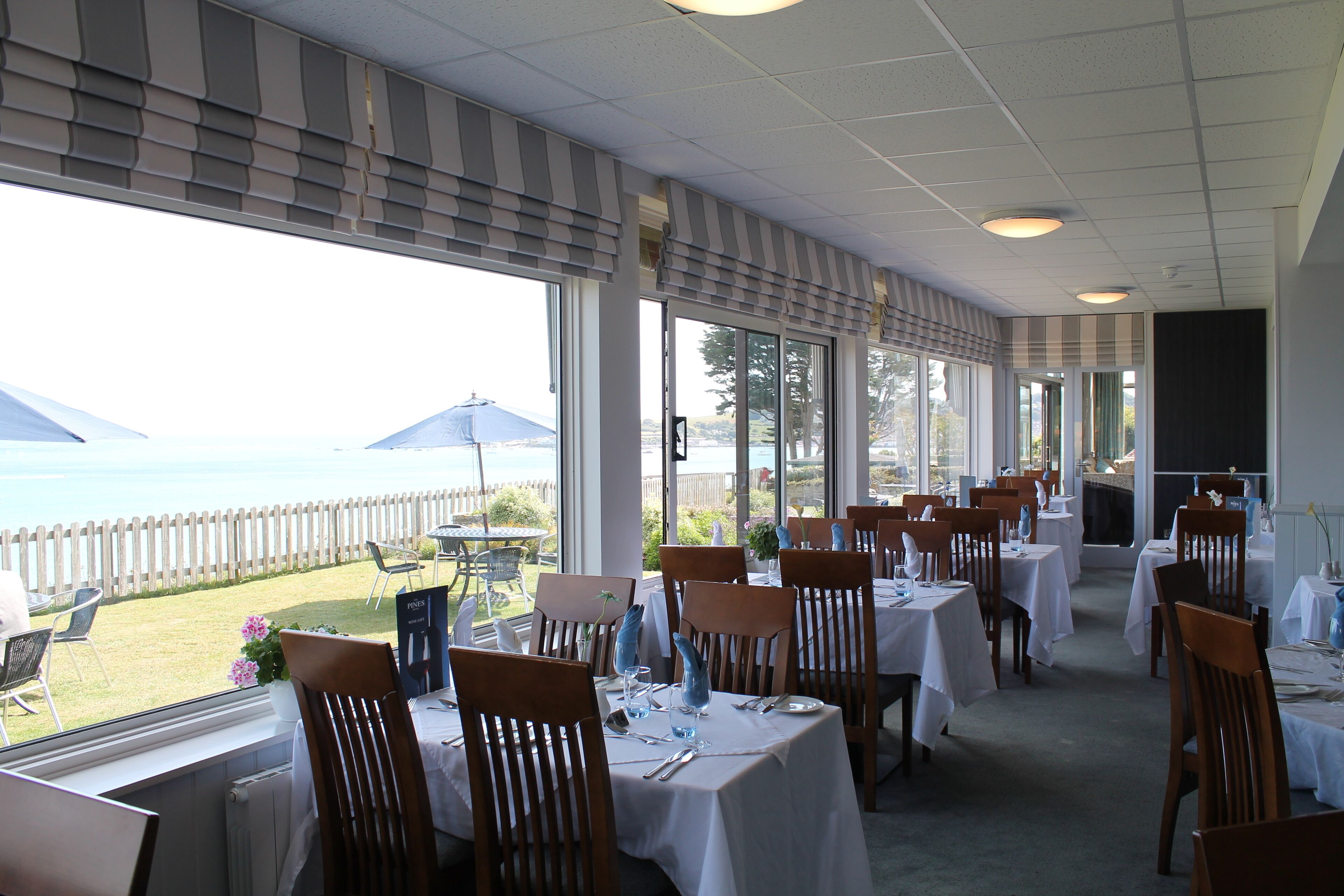A restaurant with tables and chairs arranged for dining, large windows with a view of the sea, and a covered outdoor seating area.