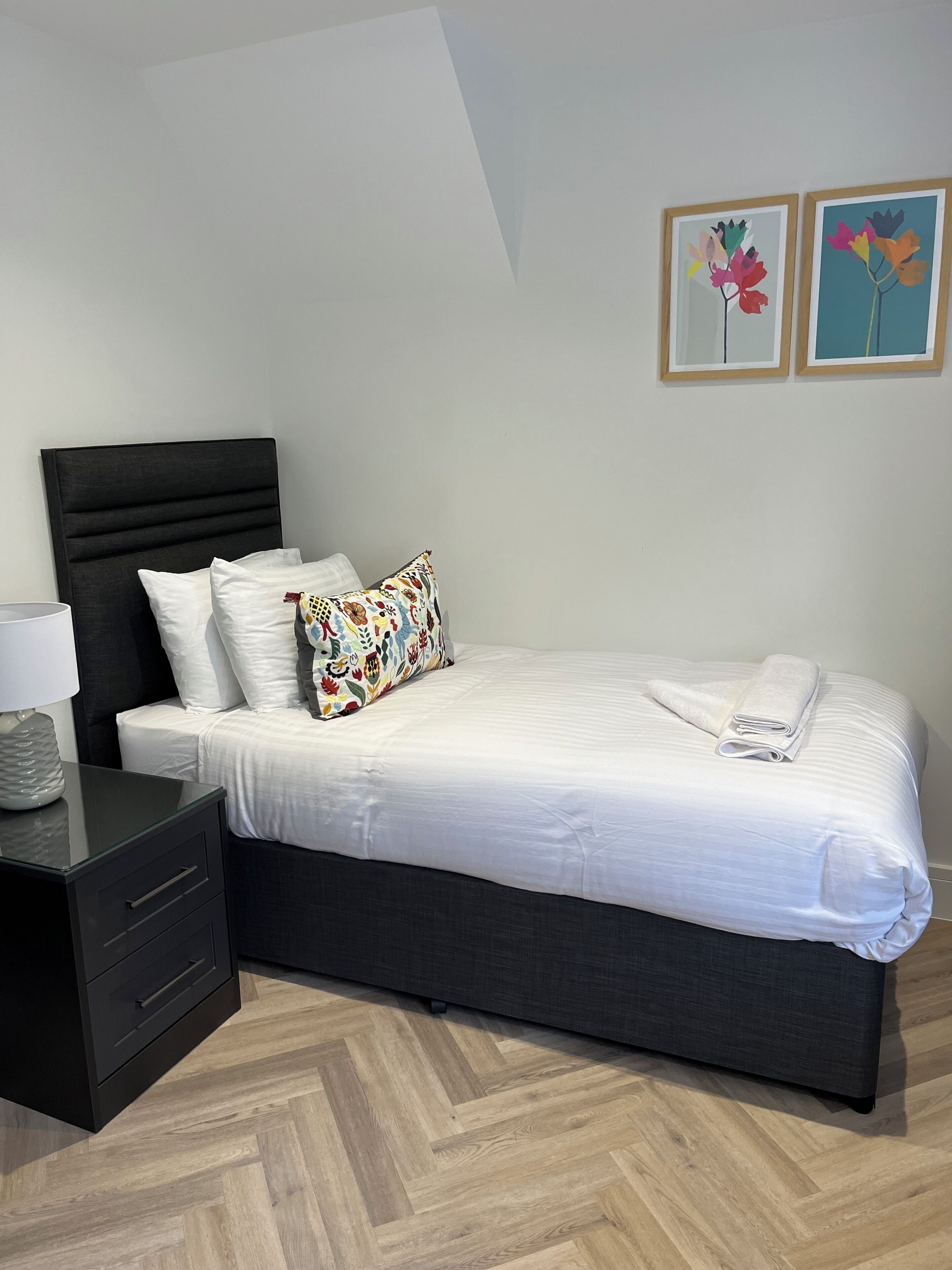 A neatly made bed with white linens and a colorful pillow, a black nightstand with a lamp, and a herringbone-patterned wooden floor.
