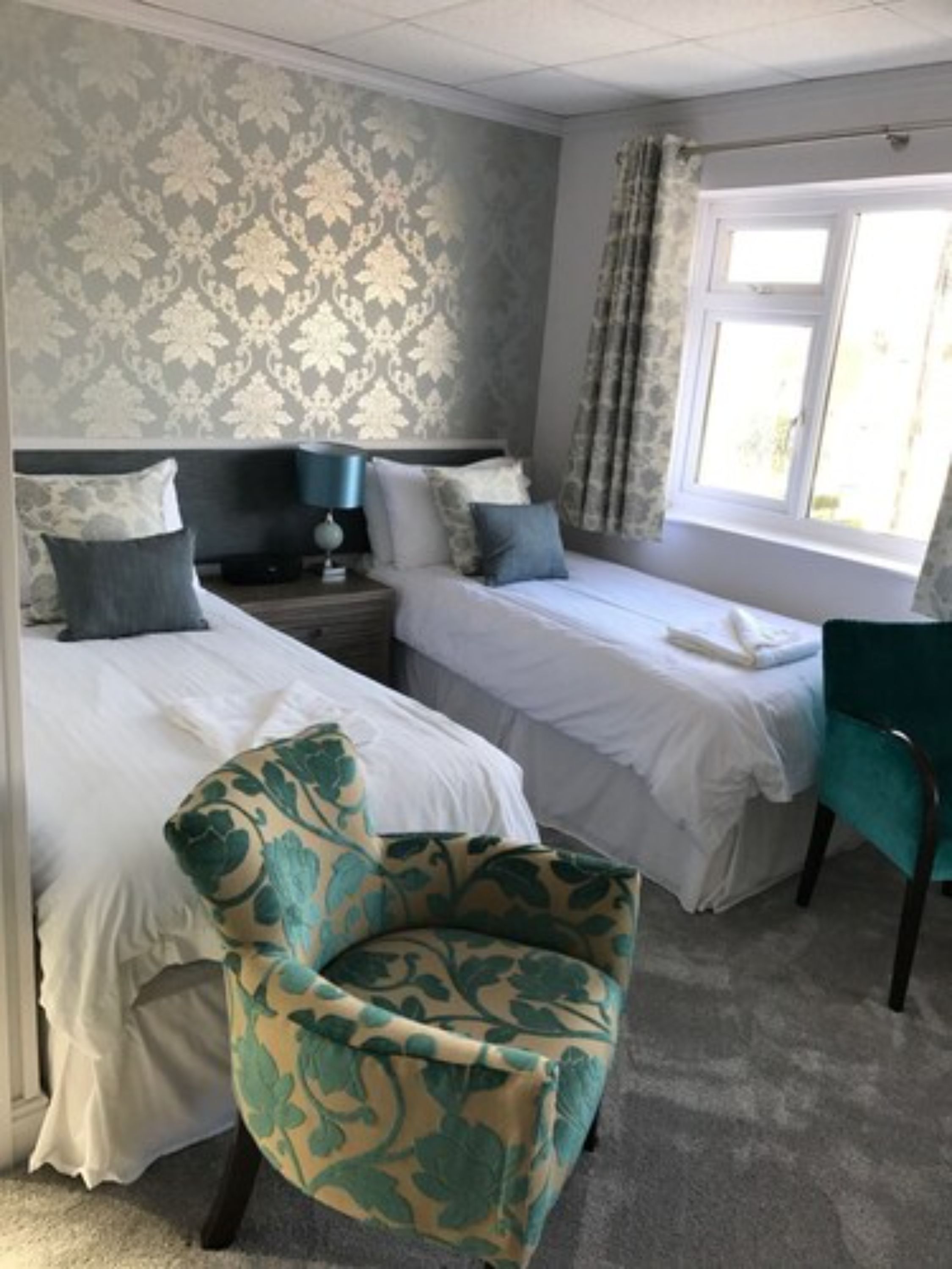 Une chambre d’hôtel avec deux lits simples, une chaise, une table de chevet et une fenêtre avec des rideaux.