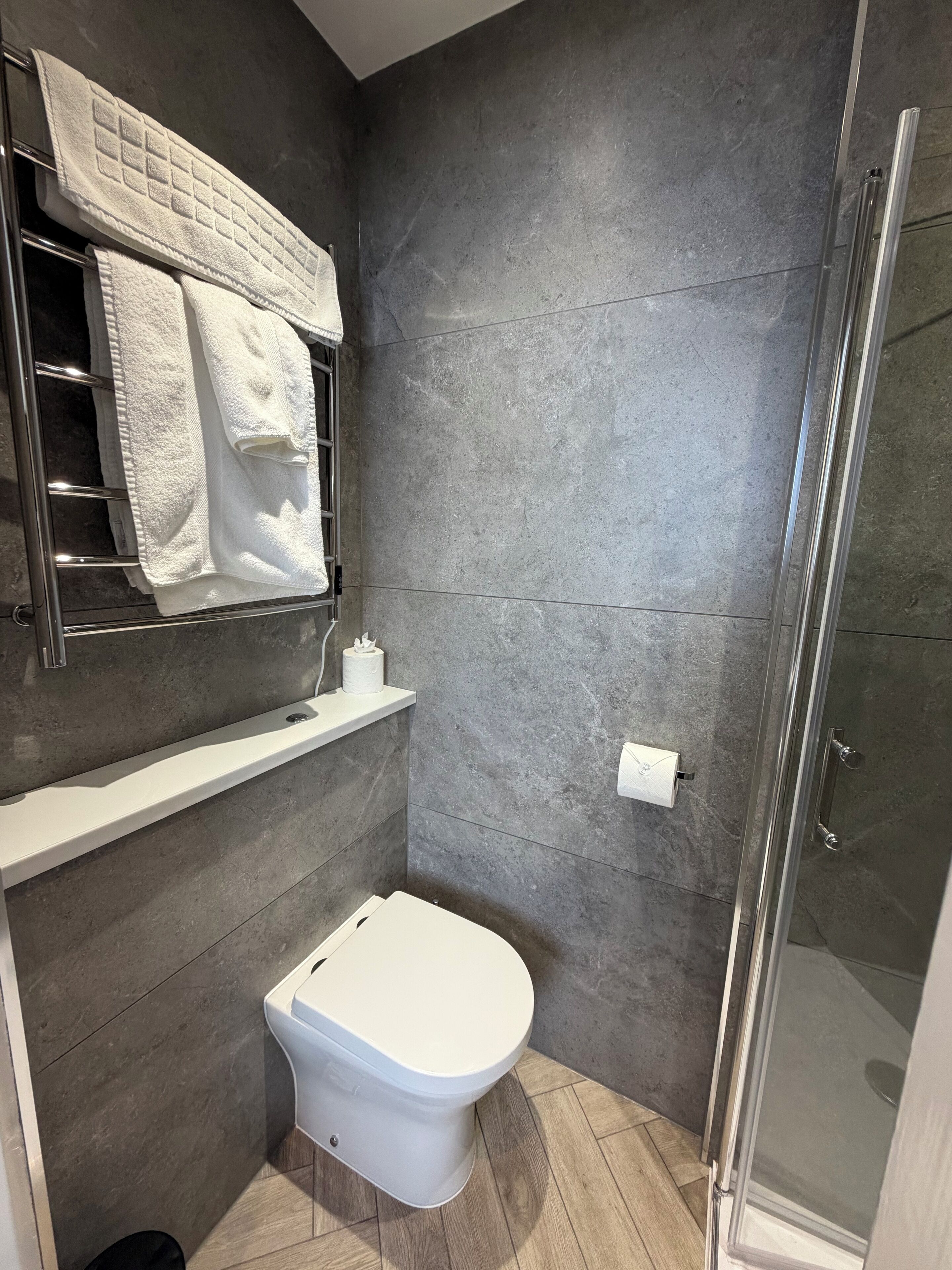 Un baño moderno con inodoro, toallero con toallas, repisa con dispensador de papel higiénico y zona de ducha.