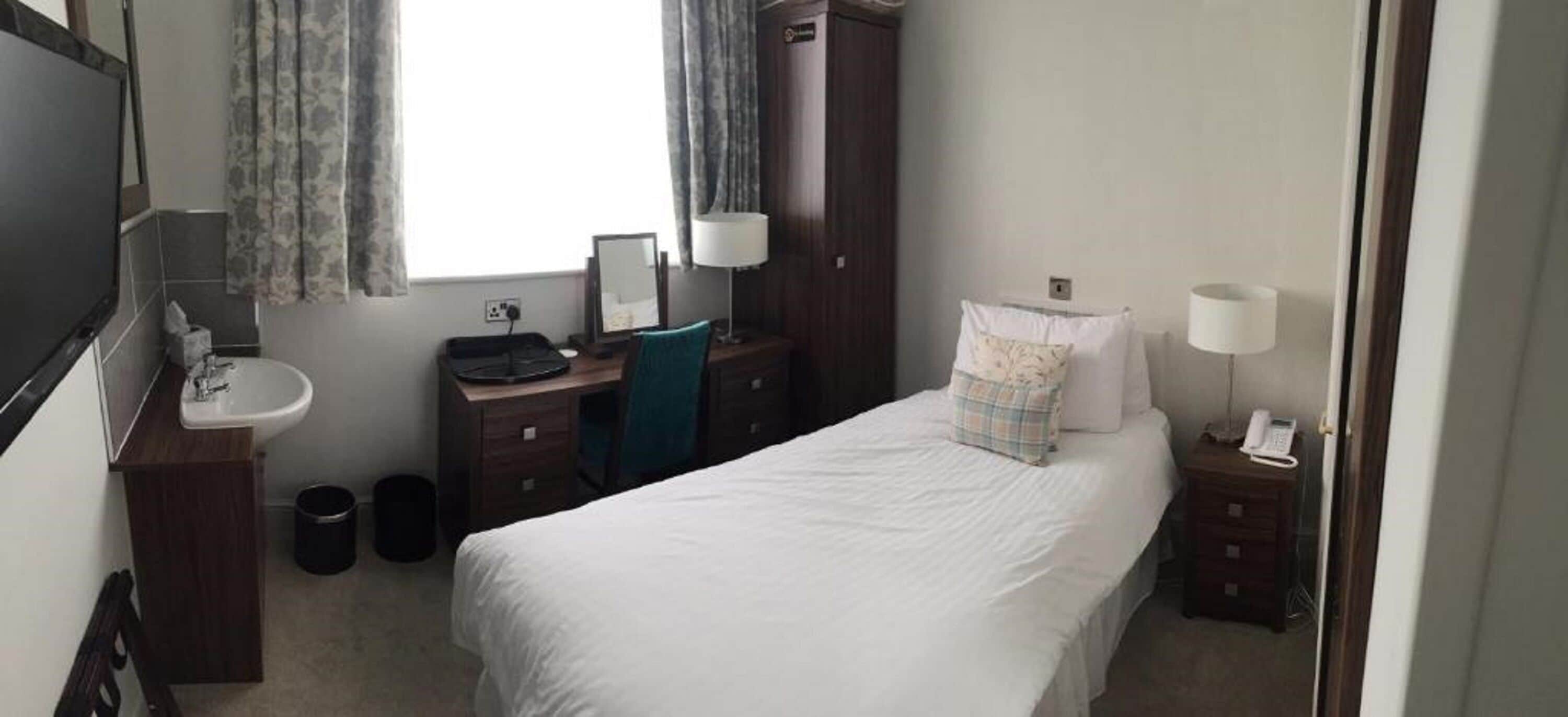 A hotel room with a bed, a desk with a computer, a chair, a bedside table with a lamp, a telephone, and a window with curtains.
