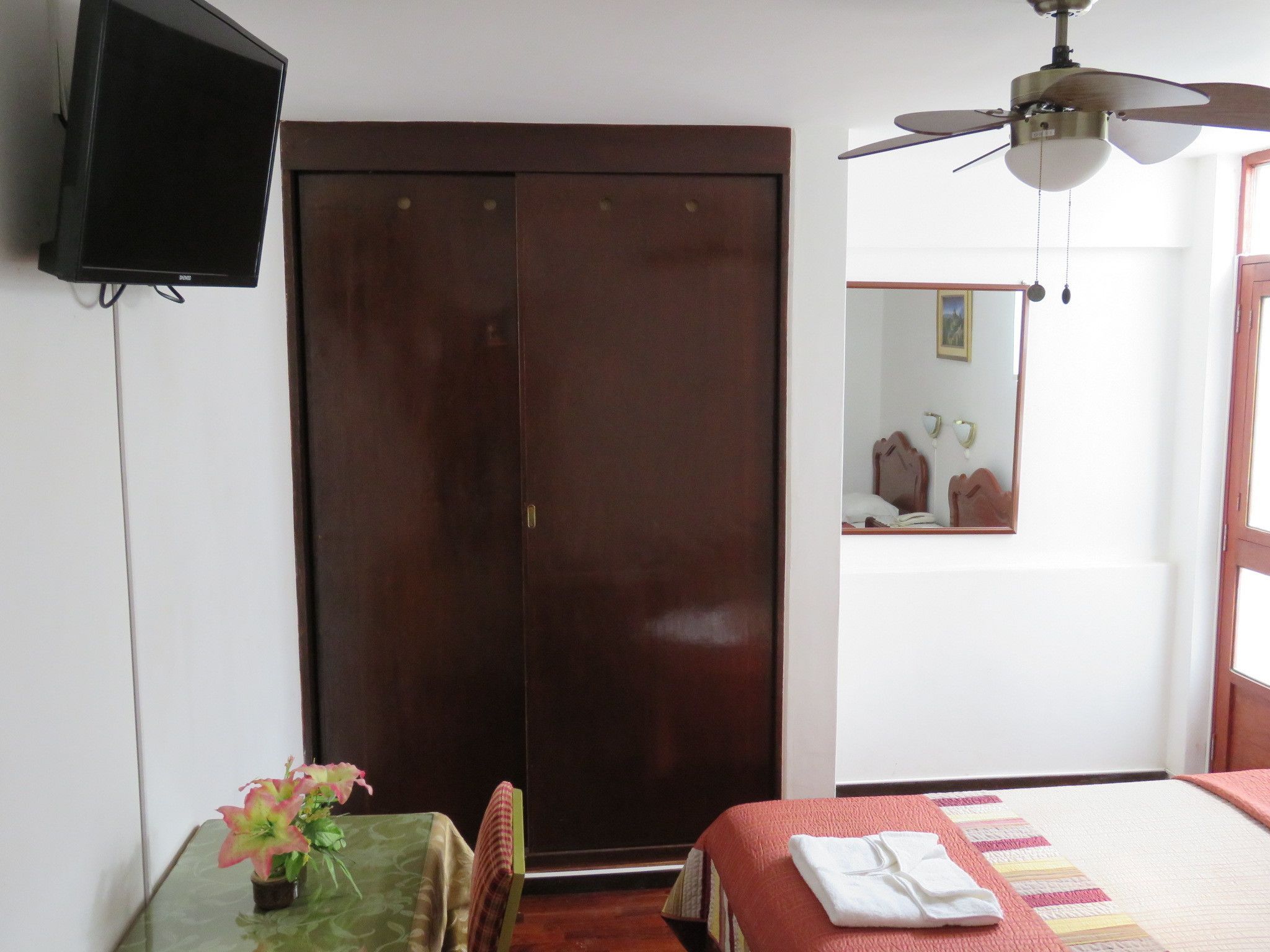 A bedroom with a bed, a wooden wardrobe, a ceiling fan, a TV mounted on the wall, and a small table with a plant.