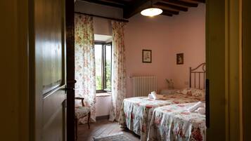 Un dormitorio con cama, una silla, una ventana con cortinas, un radiador y un cuadro en la pared.