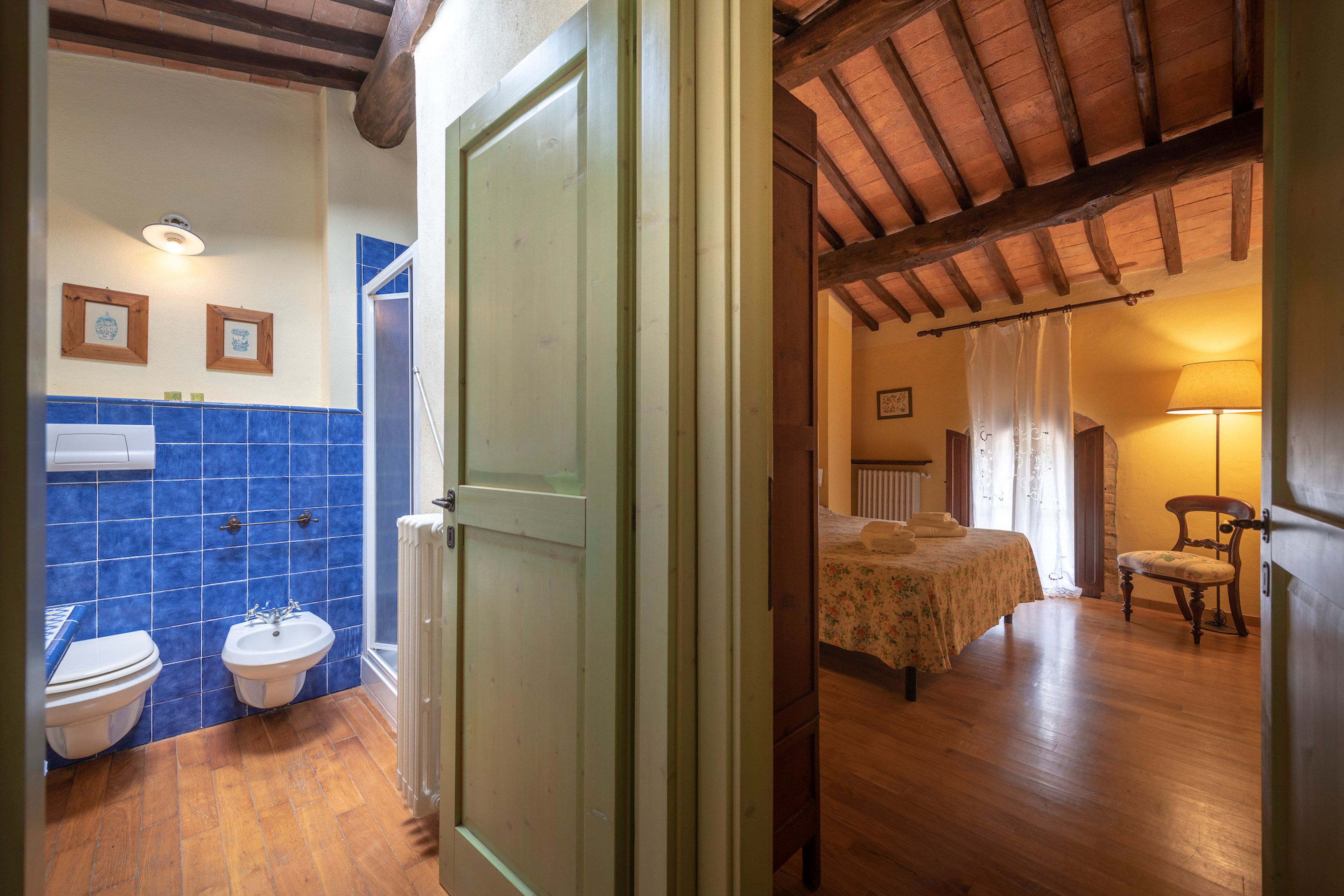 A bathroom with a toilet, bidet, and sink, a bedroom with a bed and chair, and a wooden ceiling.