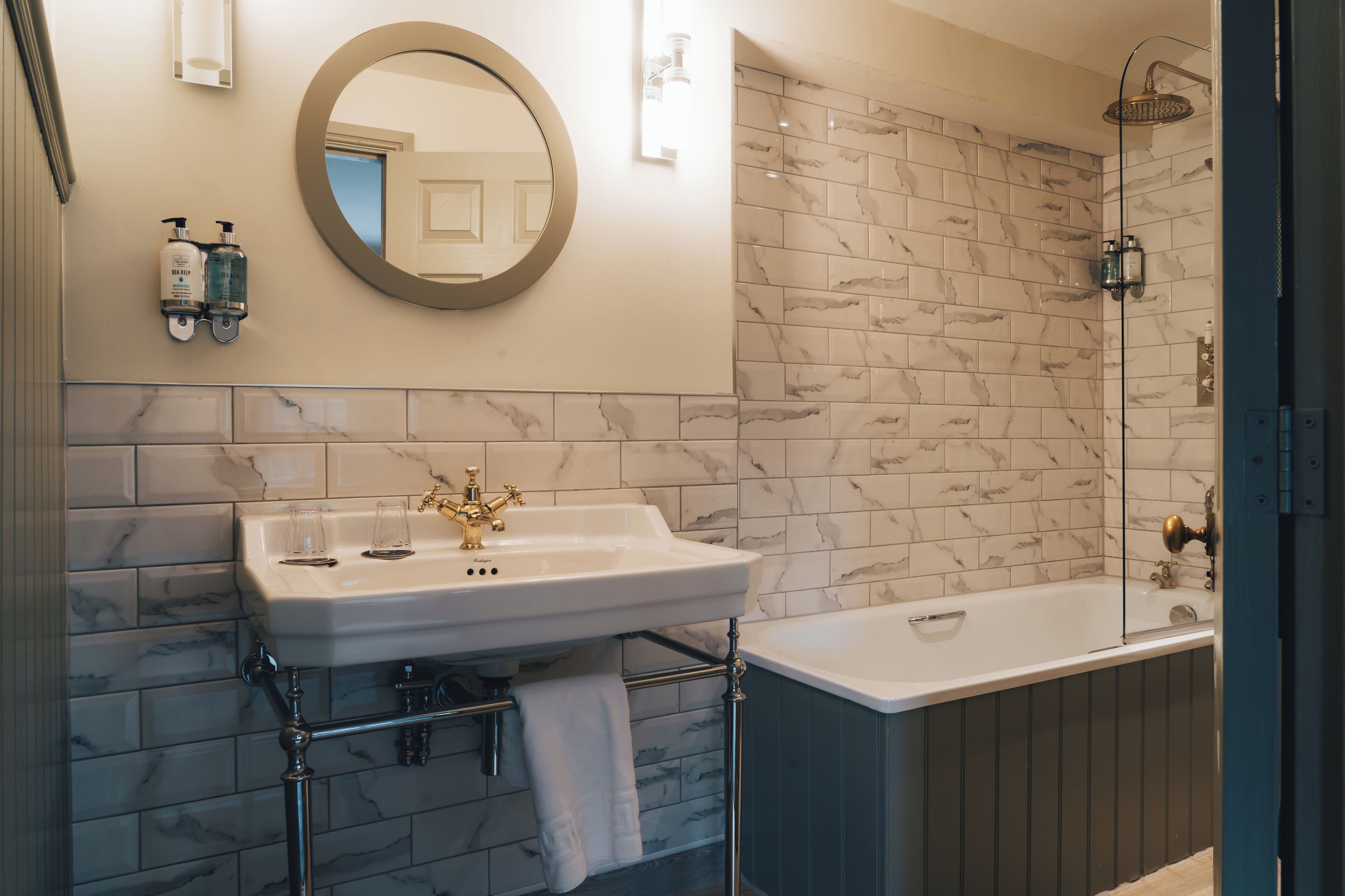 A bathroom with a marble-tiled wall, a round mirror, a white sink with gold fixtures, a bathtub, and a shower with a glass enclosure.