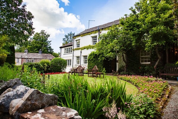 A white building with ivy, surrounded by a well-maintained garden with flowers and a lawn.