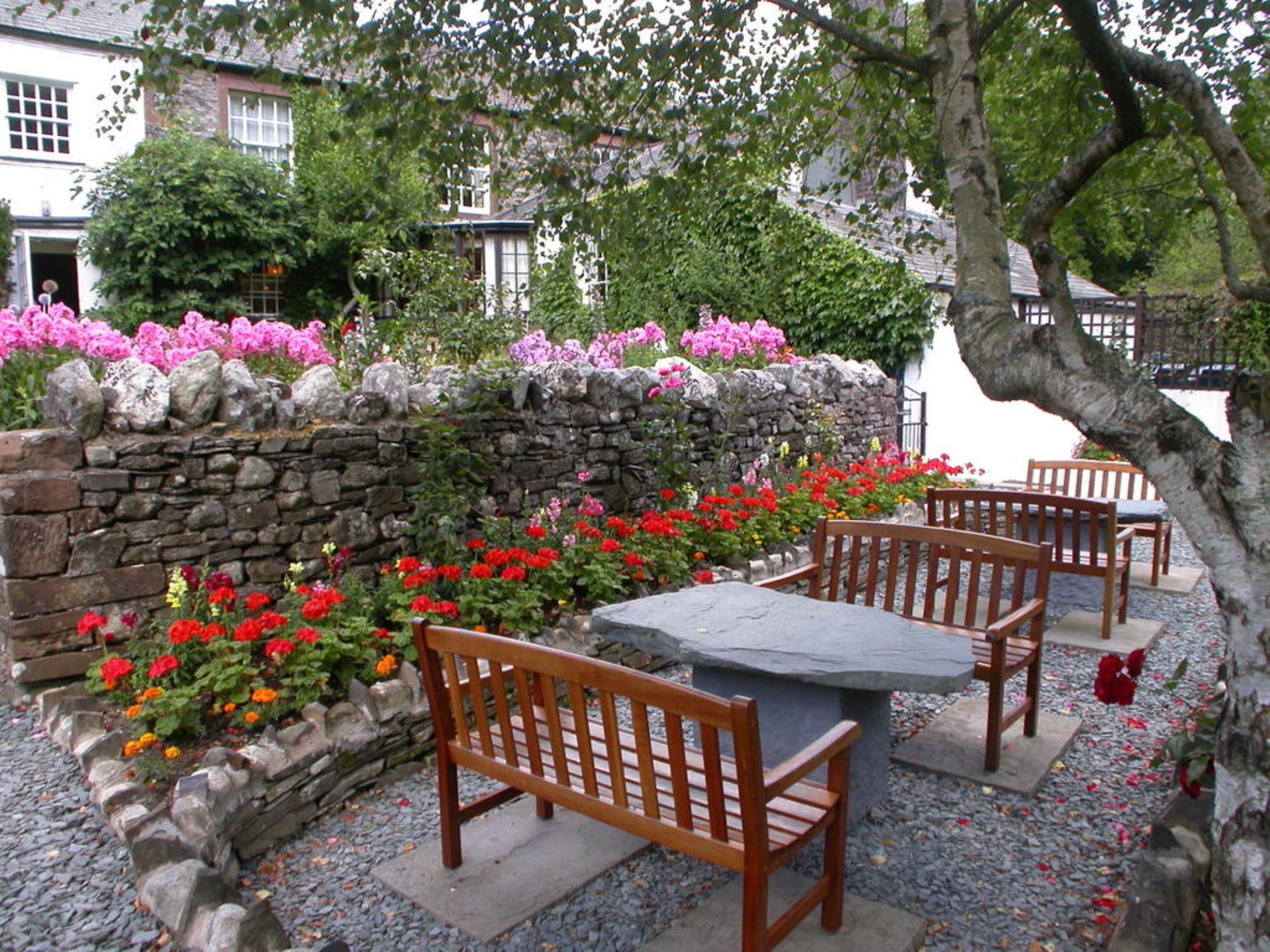 A stone wall with a bench and table in front, surrounded by colorful flowers and a stone pathway.
