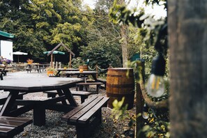 An outdoor dining area with wooden picnic tables and benches, surrounded by trees and a rustic building.
