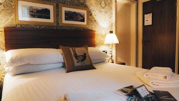 A neatly made bed with white linens, a wooden headboard, and a decorative pillow. There is a folded towel on the bed and a framed picture above it.
