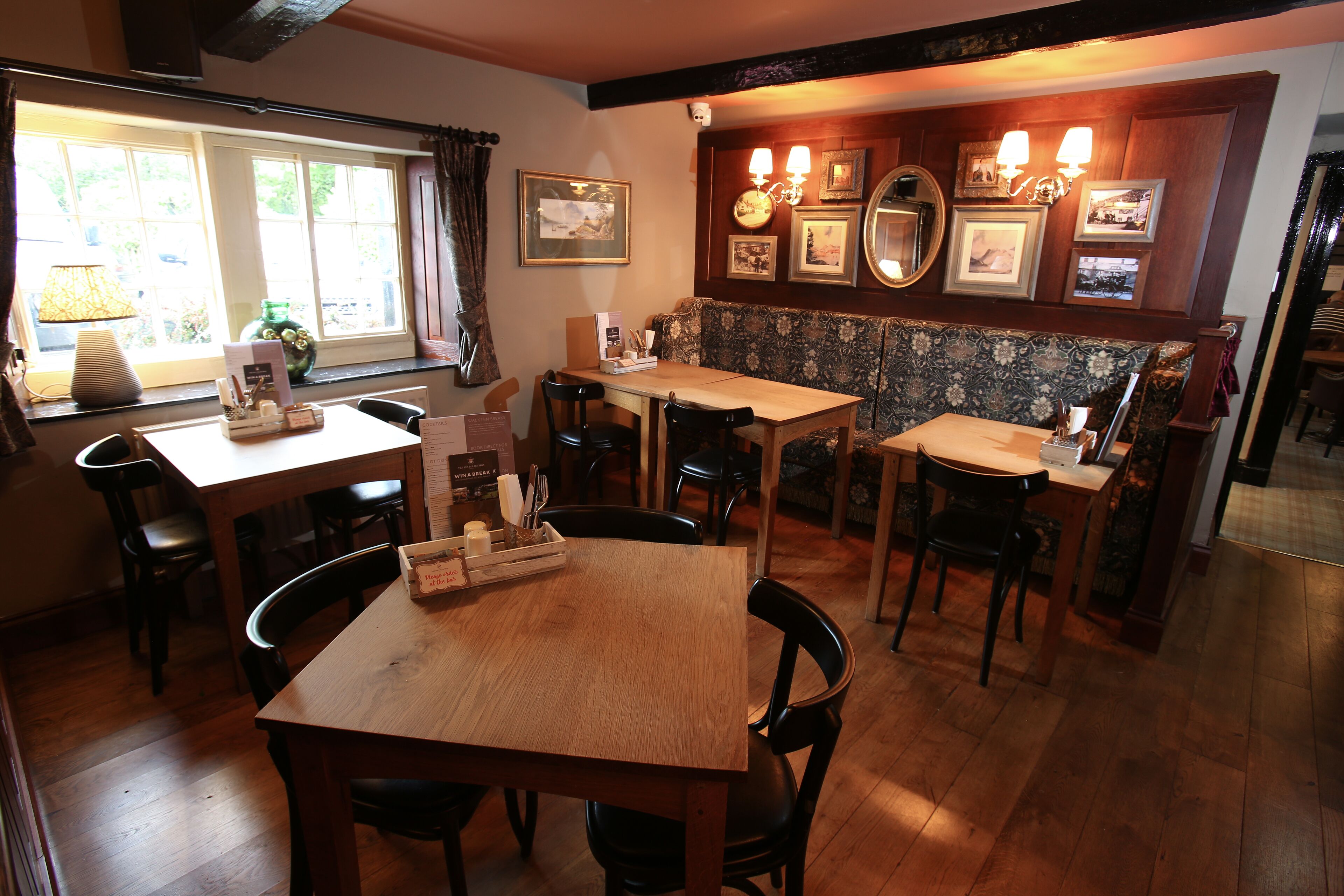 A dining area with wooden tables and chairs, a window with curtains, and wall-mounted picture frames and lights.