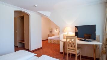 Quarto de hotel com cama, escrivaninha, cadeira, televisão e banheiro pequeno.