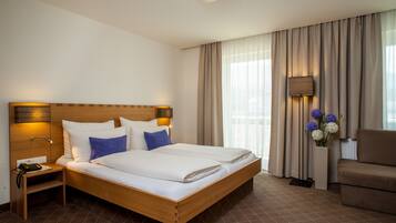 Quarto de hotel com cama de madeira, mesa de cabeceira, luminária, cadeira e vista para o exterior através de amplas janelas.