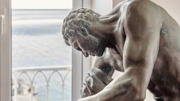 Una estatua de un hombre sentado con expresión pensativa, ubicada en una habitación con vista al mar a través de una ventana.