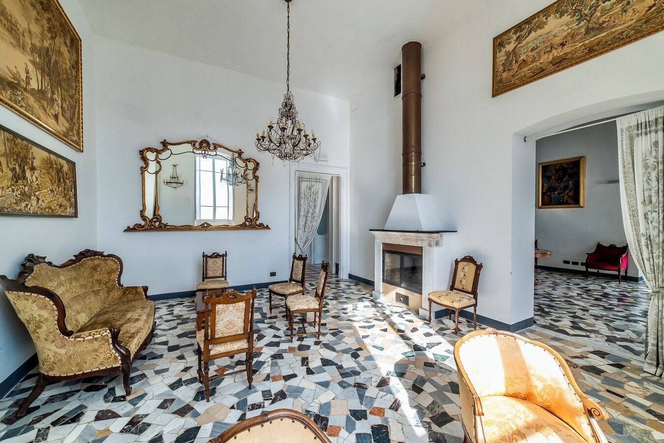 A room with a fireplace, ornate chairs, a chandelier, and patterned flooring.