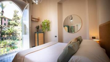 Un dormitorio con una cama grande, un espejo redondo, una mesita de noche con una planta y vistas a un jardín a través de una ventana.