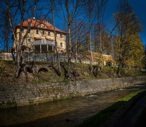 A historic building with a red roof and a balcony, located beside a river with a stone wall and trees. - Rezydencja Grawert - Victorian Boutique & SPA (Ladek-Zdroj)