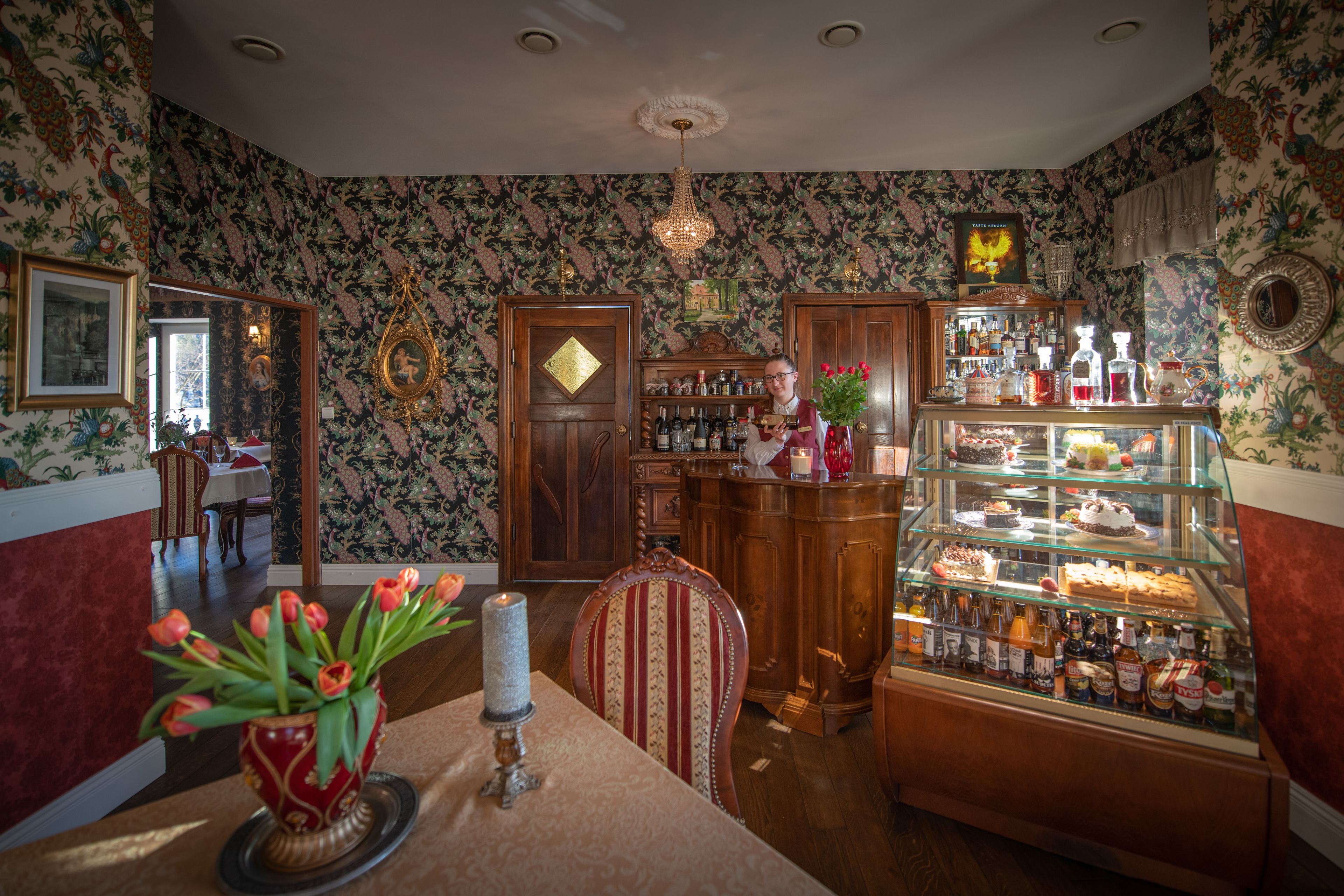 Una habitación con papel tapiz floral, un mueble de madera con licores, una vitrina de vidrio con pasteles y un jarrón rojo con tulipanes.