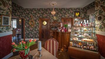 Una habitación con papel tapiz floral, un mueble de madera con licores, una vitrina de vidrio con pasteles y un jarrón rojo con tulipanes.