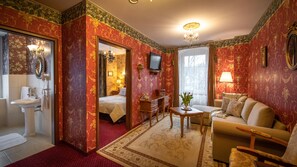 A room with a bed, a sitting area with a sofa and armchair, a small table, a television, and a bathroom with a sink and mirror. - Rezydencja Grawert - Victorian Boutique & SPA (Ladek-Zdroj)