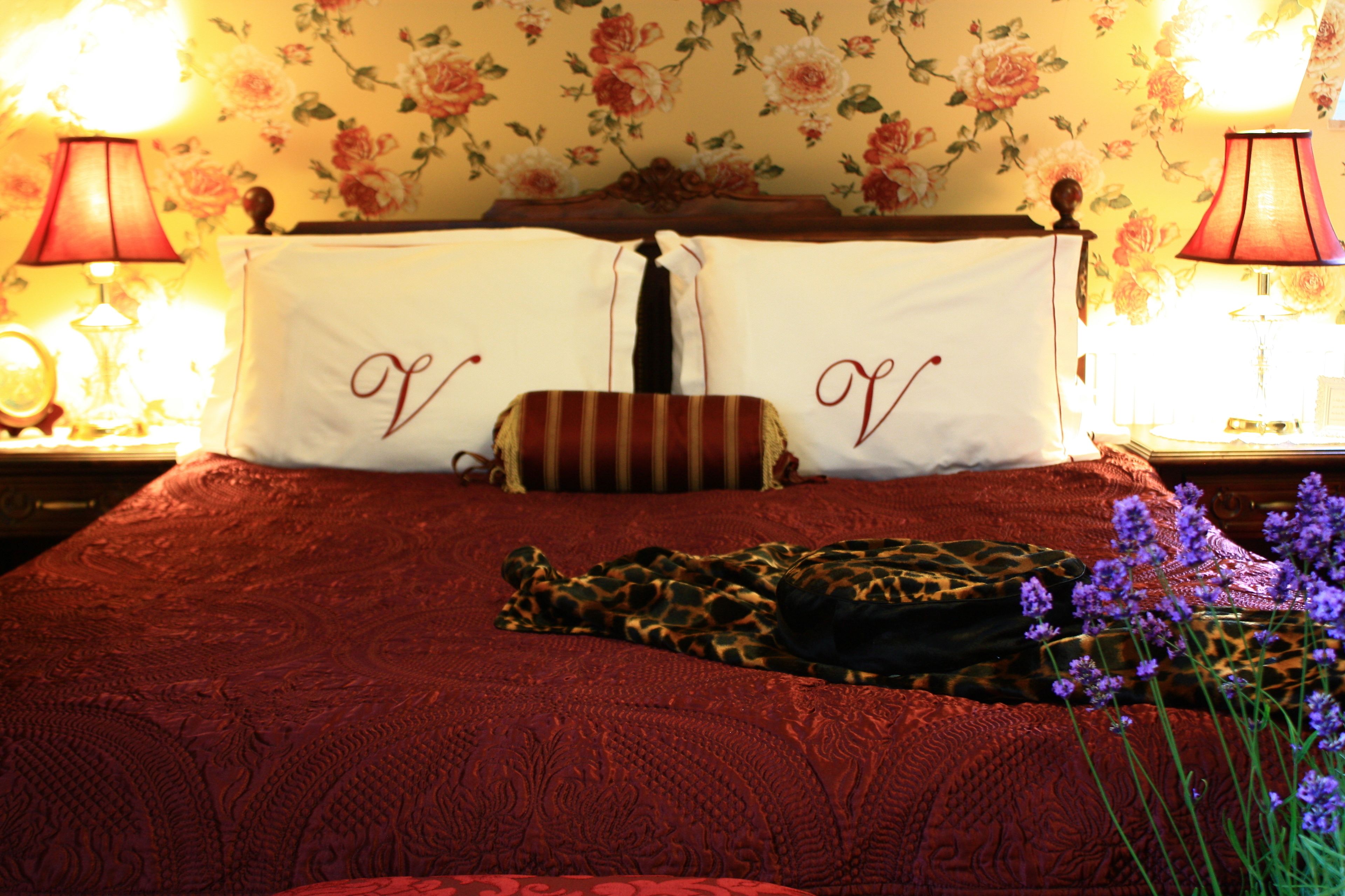 Una cama con un edredón rojo, dos almohadas con un monograma y una almohada de estampado de leopardo, además de un florero con flores moradas.