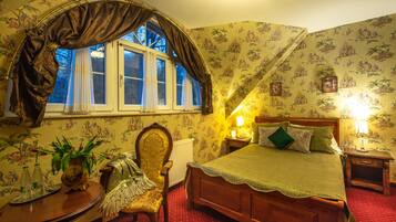 A bedroom with a large bed, a wooden chair, a bedside table, a window with curtains, and floral wallpaper.