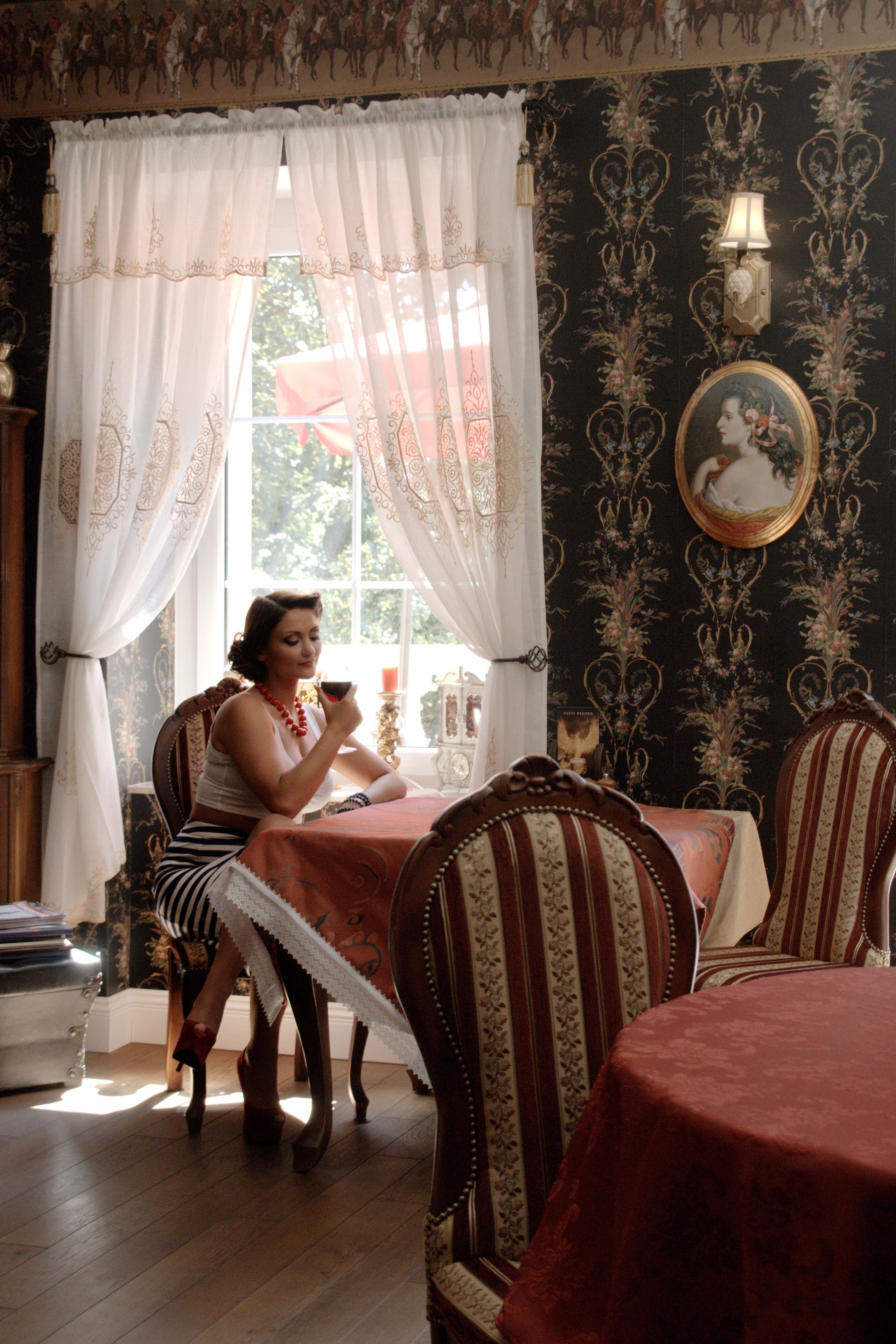 A room with a dining table, chairs, a window with curtains, a lamp, and a framed picture on the wall.