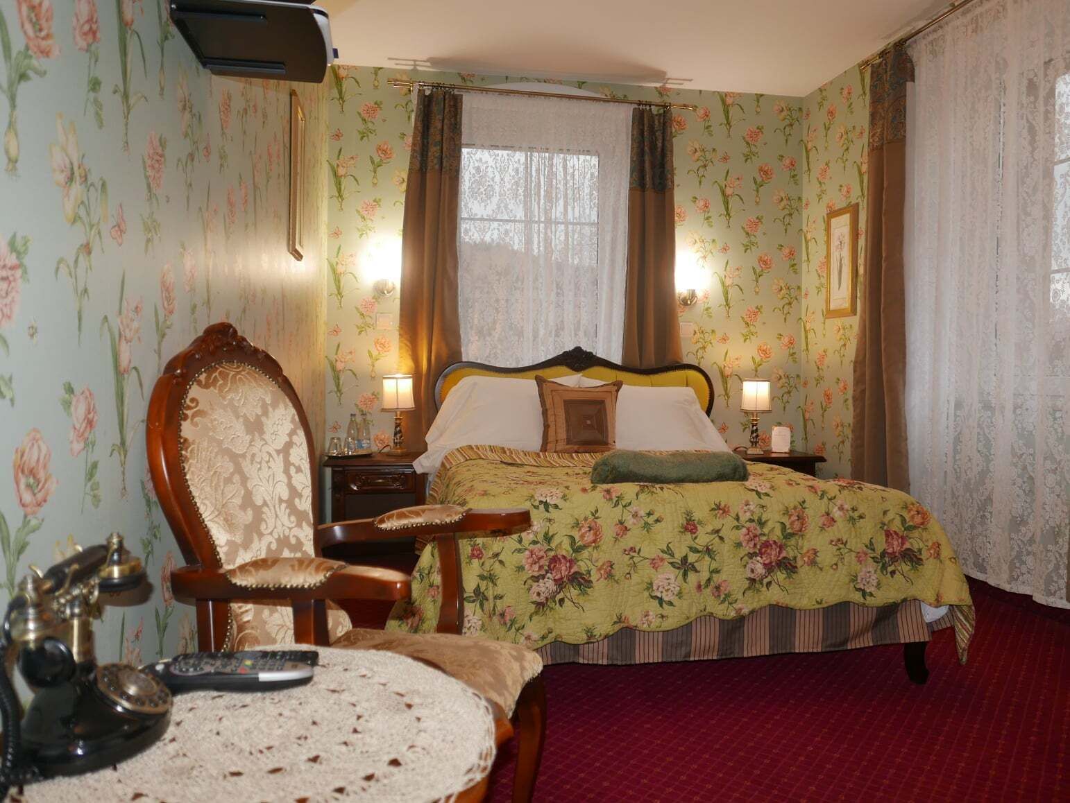 Une chambre à coucher avec un papier peint à motifs floraux, un lit recouvert d’une couvre-lit à motifs floraux, une chaise en bois, une table de chevet avec une lampe et un téléphone.