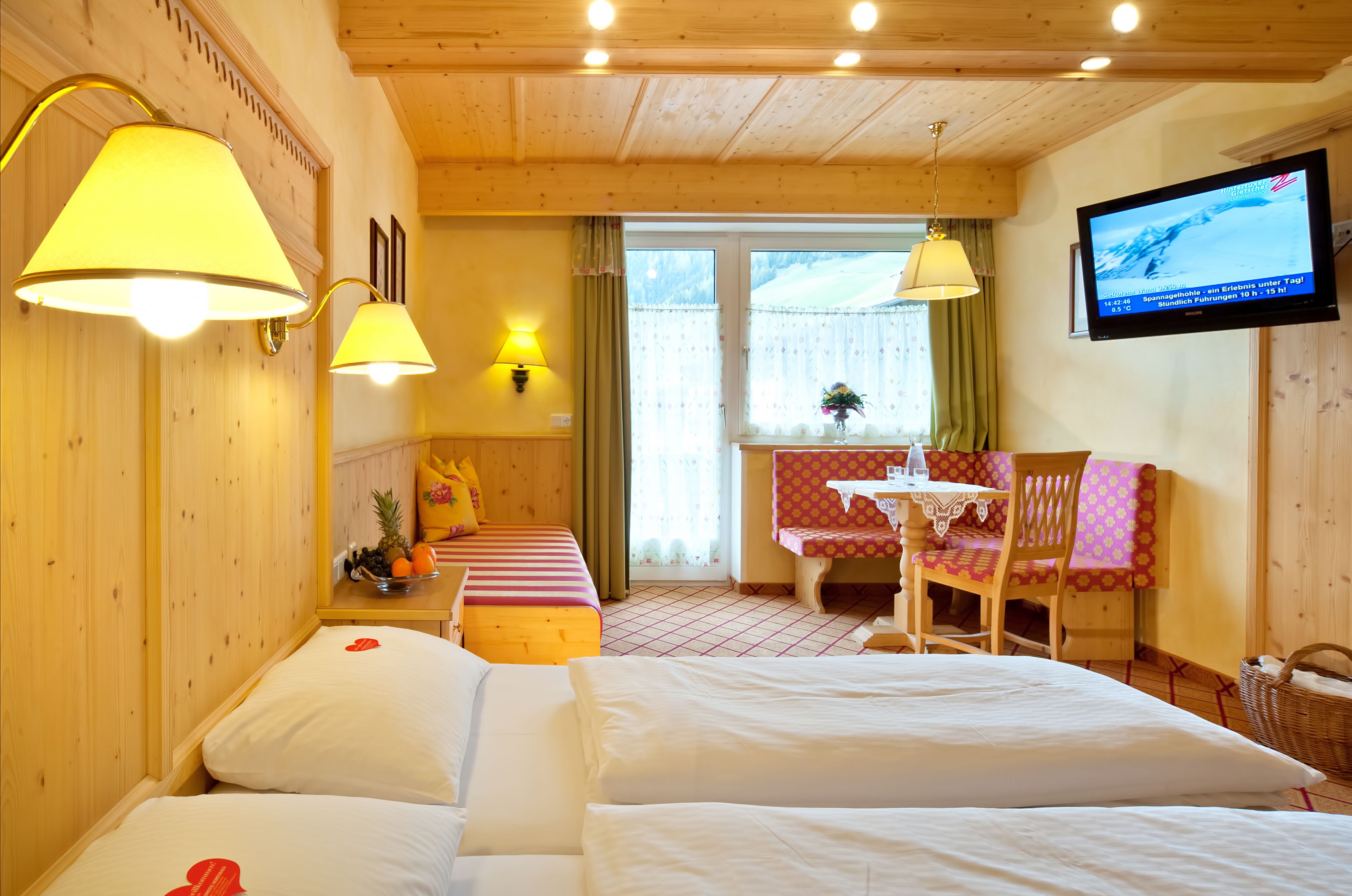 A hotel room with wooden walls and ceiling, a bed with white linens, a small table with a pink tablecloth, and a television mounted on the wall.