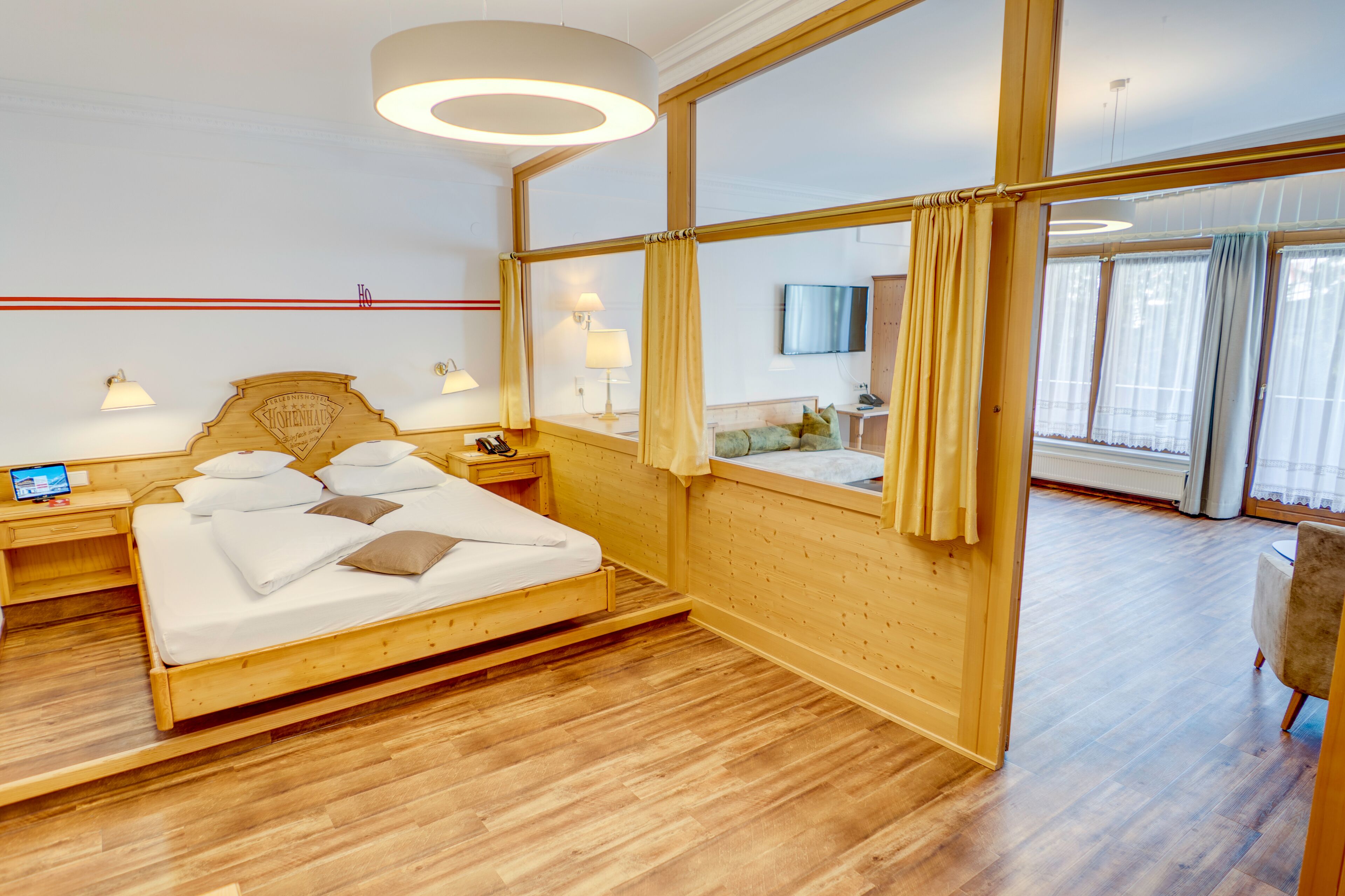 Habitación de hotel con cama de madera, lámparas de noche, televisor de pantalla plana y un amplio ventanal con cortinas.