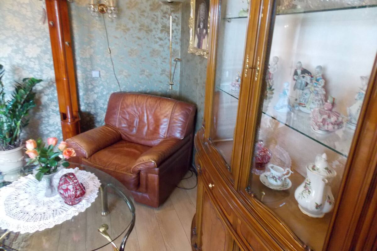 A room with a brown leather armchair, a glass table with a lace doily, a potted plant, and a glass cabinet with decorative items.