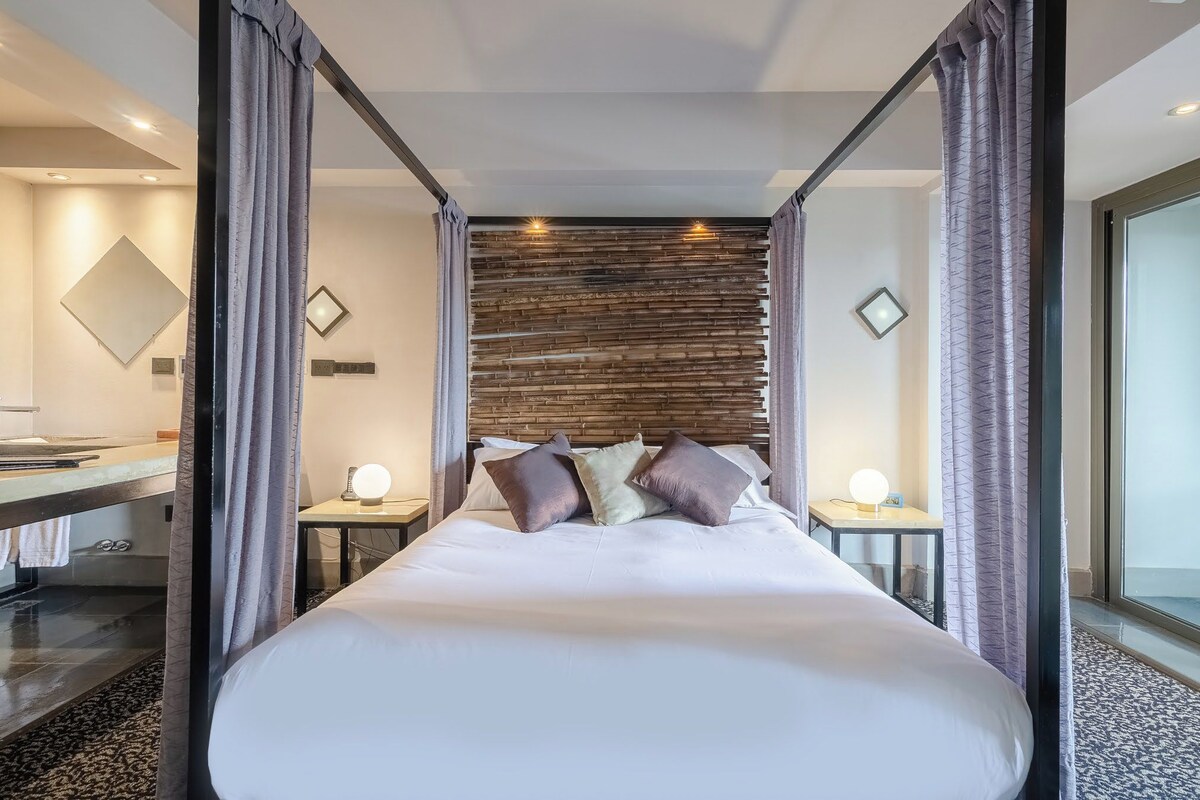 A modern hotel room with a large bed, two bedside tables with lamps, a wooden headboard, and a large mirror on the wall.