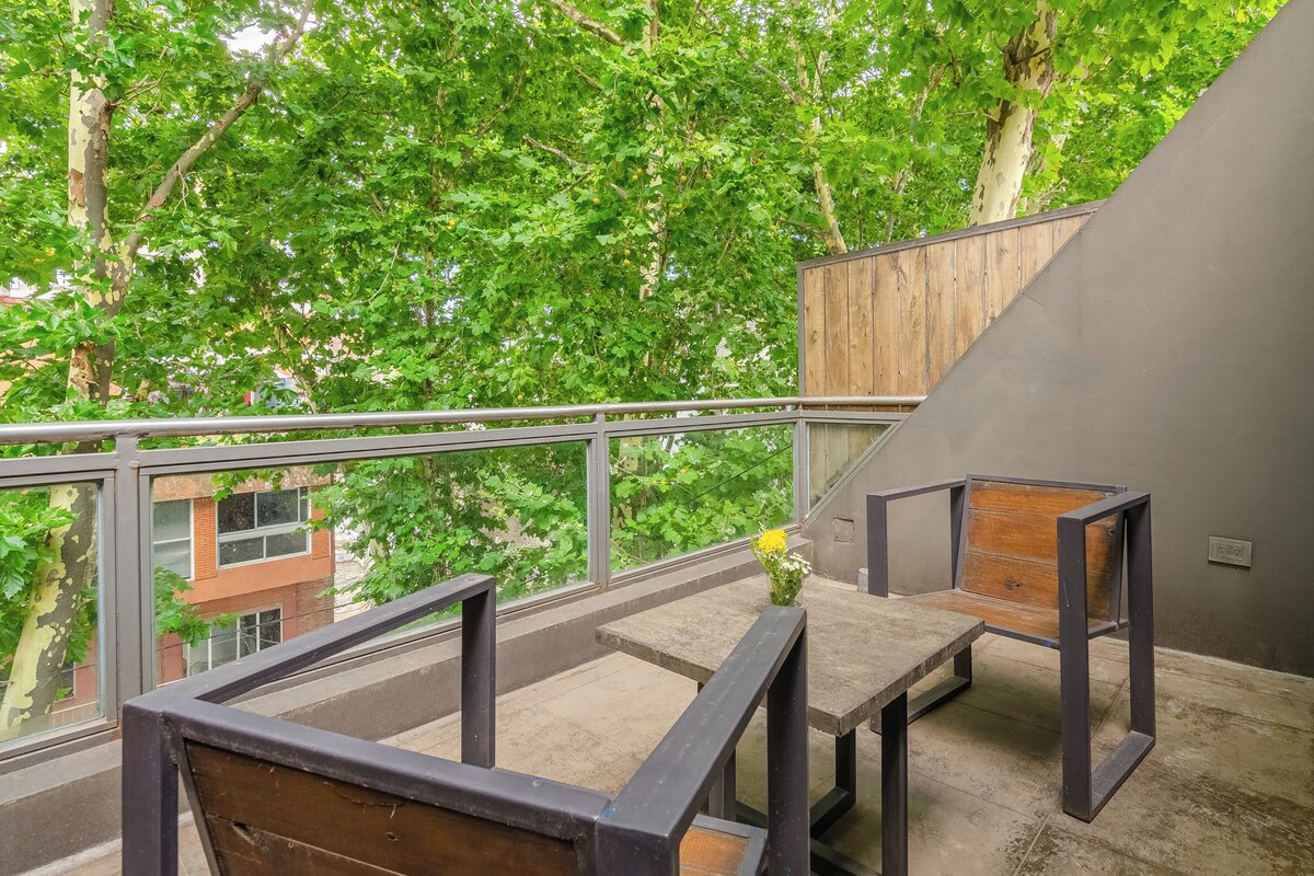 Un balcon avec vue sur des arbres et un immeuble. On y trouve deux bancs en bois et une rambarde en métal.