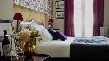 Una habitación de hotel con una cama, una mesita de noche con una botella de vino y copas, un florero con flores y una ventana con cortinas.