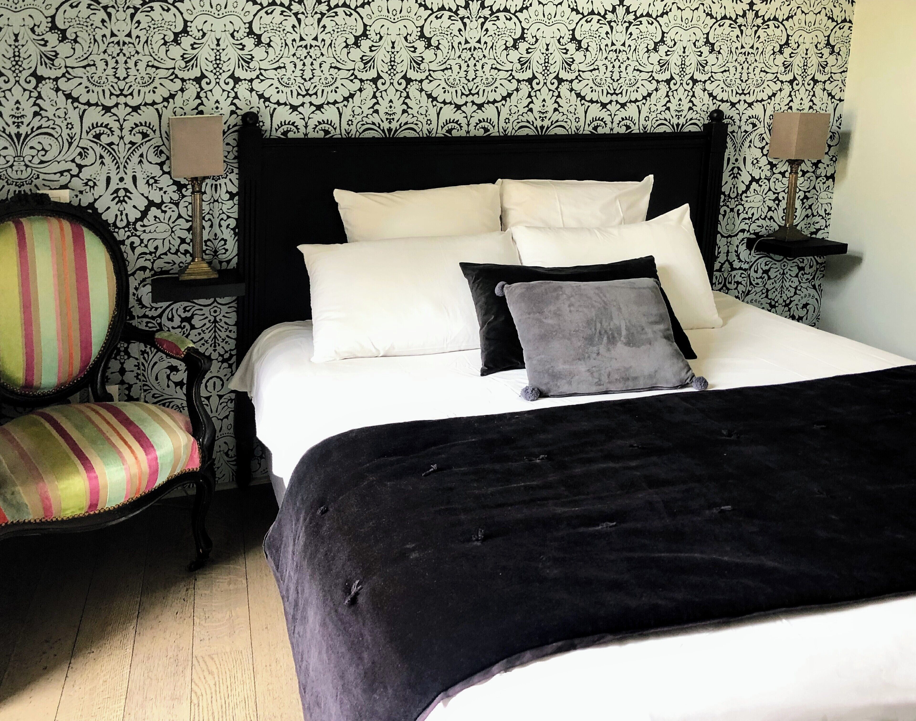 Een net opgemaakt bed met witte en grijze kussens, een zwart hoofdbord en een grijze plaid. Er staat een gestreept fauteuil met kussen en een klein tafeltje met lamp naast het bed.