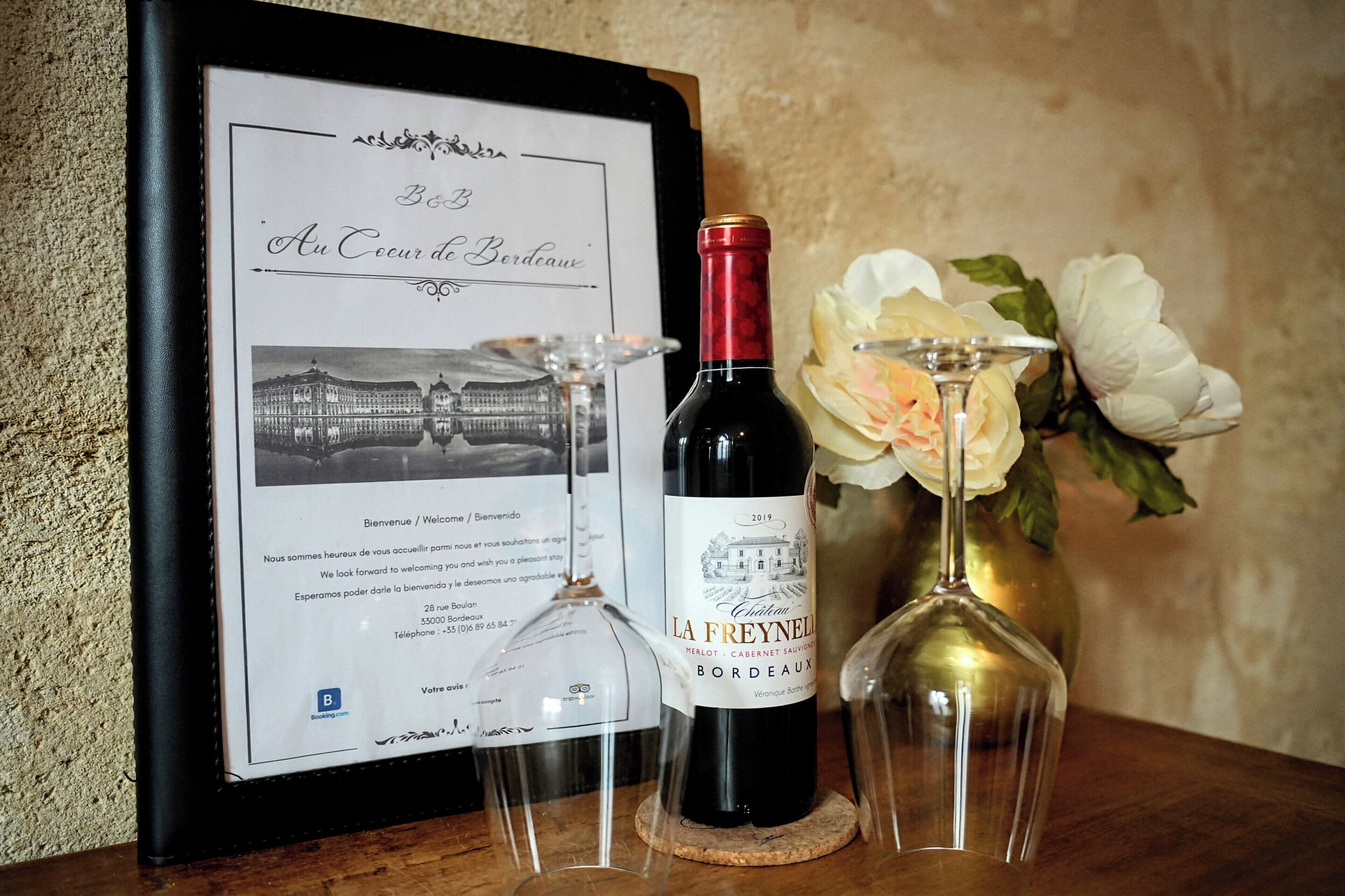 A framed welcome sign with a building illustration, a bottle of La Freyella wine, and two wine glasses on a wooden table.