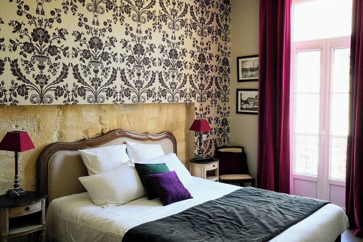 Un lit bien fait, agrémenté d’oreillers blancs et violets, d’une couvre-lit sombre et d’une tête de lit en bois. Deux tables de chevet avec des lampes se trouvent de part et d’autre du lit, dont l’une est surmontée d’un abat-jour rouge. La pièce est décorée d’un papier peint à motifs floraux et possède une fenêtre avec des rideaux rouges.