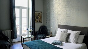 A bedroom with a large bed, a nightstand, a chair, a window with curtains, and a wall with a patterned wallpaper. - Au cœur de Bordeaux B&B (Bordeaux)