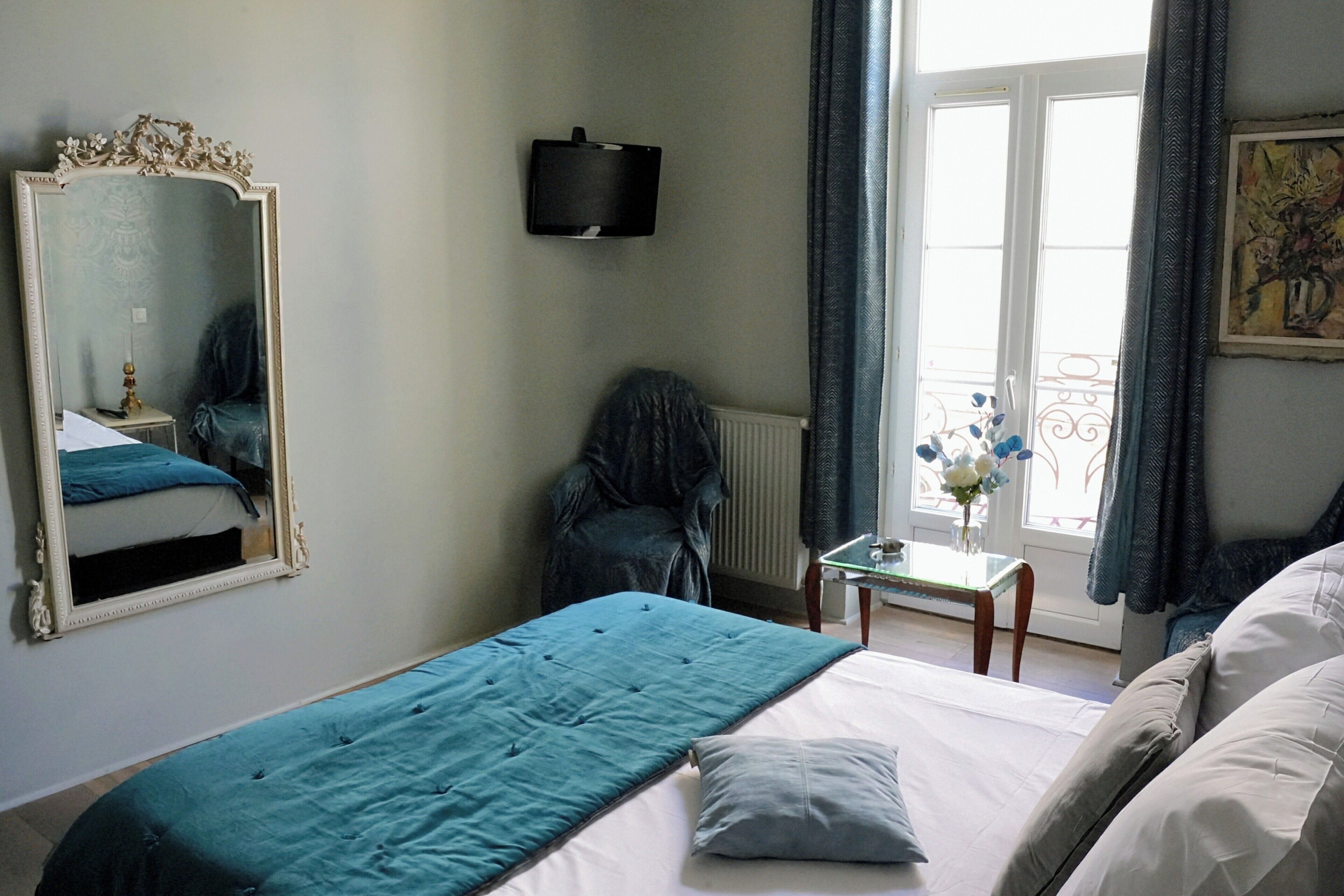 A bedroom with a large bed, a chair, a small table with a vase, a mirror, and a window with curtains.