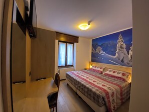 A bedroom with a bed, a desk, and a chair. There is a window with curtains and a painting of a snowy mountain scene on the wall. - Hotel Sciatori (Temu)