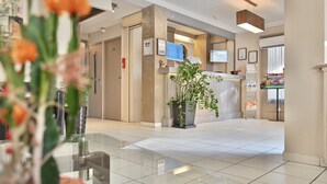 A hotel lobby with a reception desk, a large potted plant, and a tiled floor. - Hotel La Rotonde (La Ciotat)