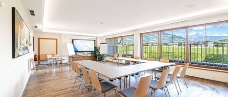 A conference room with a long table, chairs, a projector screen, and large windows offering a view of mountains.