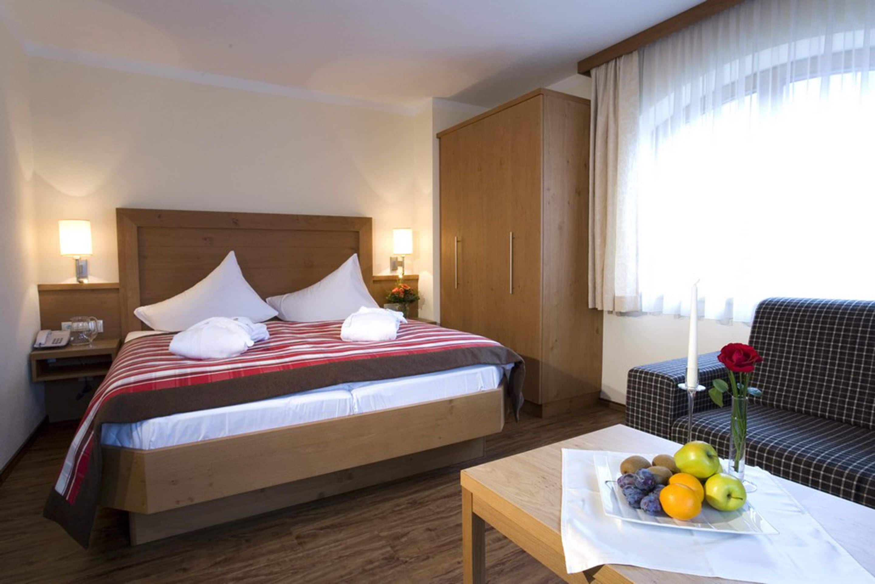 A hotel room with a bed, bedside lamps, a wooden wardrobe, a sofa, and a table with fruit and a vase with a flower.