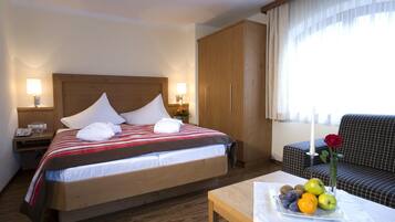 A hotel room with a bed, bedside lamps, a wooden wardrobe, a sofa, and a table with fruit and a vase with a flower.
