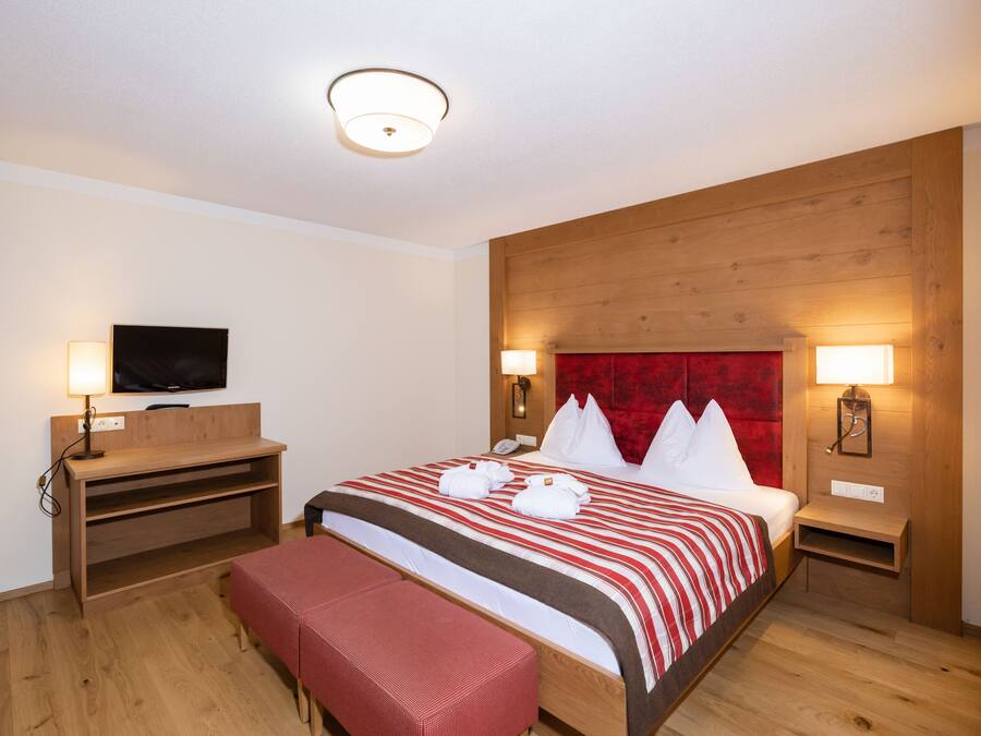 Een hotelkamer met een houten hoofdeinde, een groot bed met rood-witte beddengoed, twee nachtkastjes met lampen, een flatscreen televisie op een houten plank en een bankje voeteneinde van het bed.
