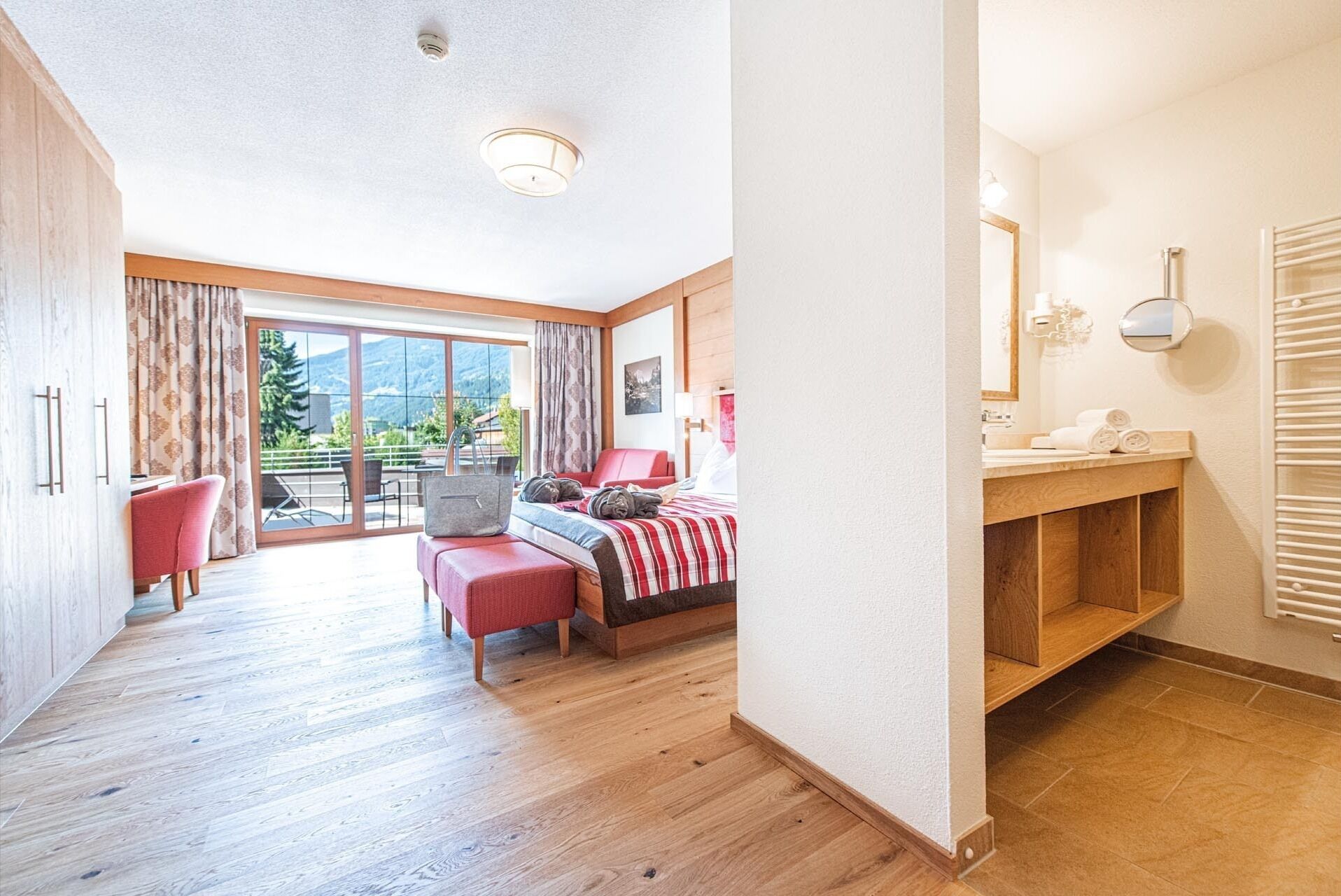 A spacious bedroom with a large sliding glass door leading to a balcony, a wooden floor, a bed with a red and white checkered bedspread, a red chair, and a small table with a handbag on it.