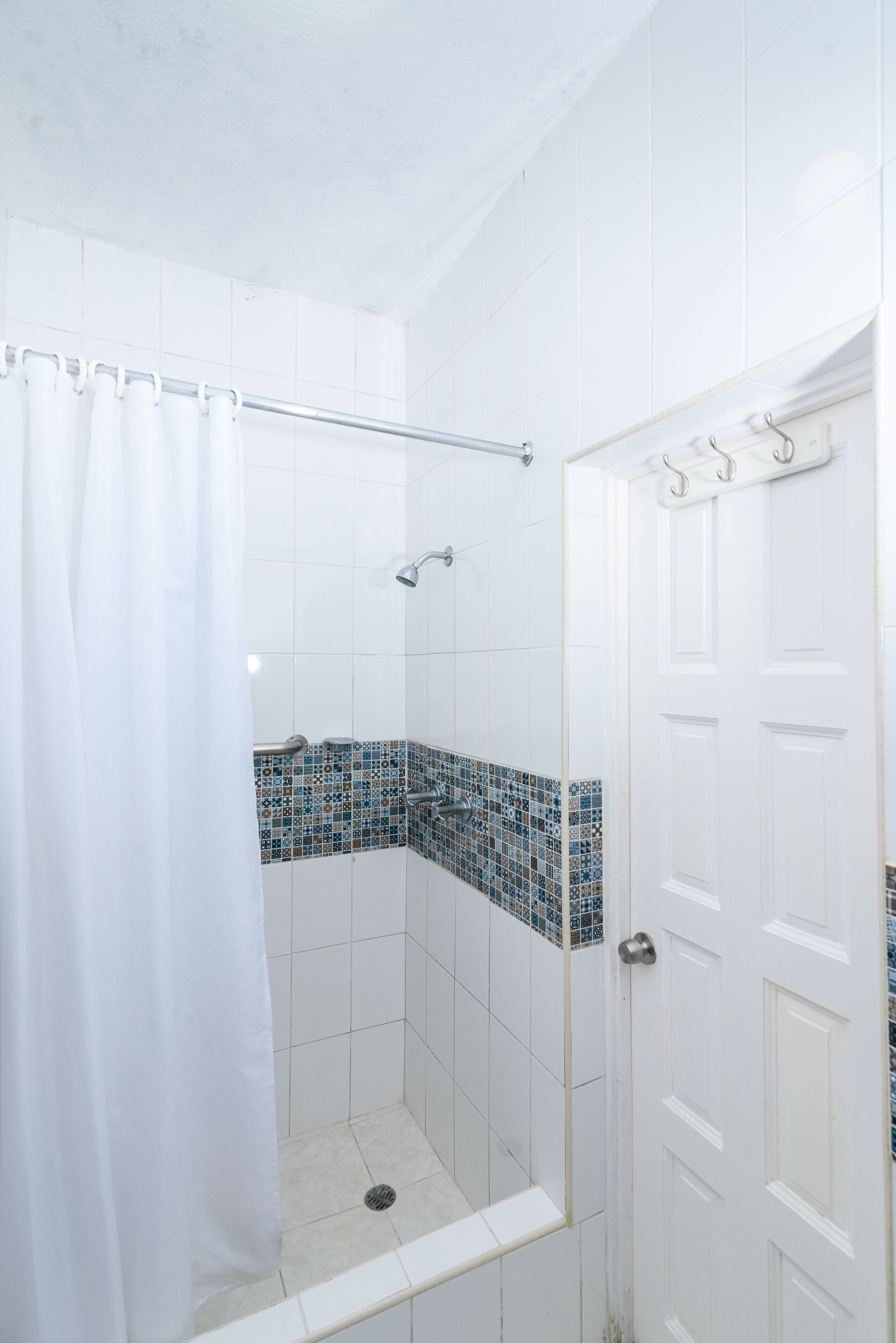 A shower with a white curtain, a tiled wall with a blue mosaic pattern, and a white door with a silver handle.