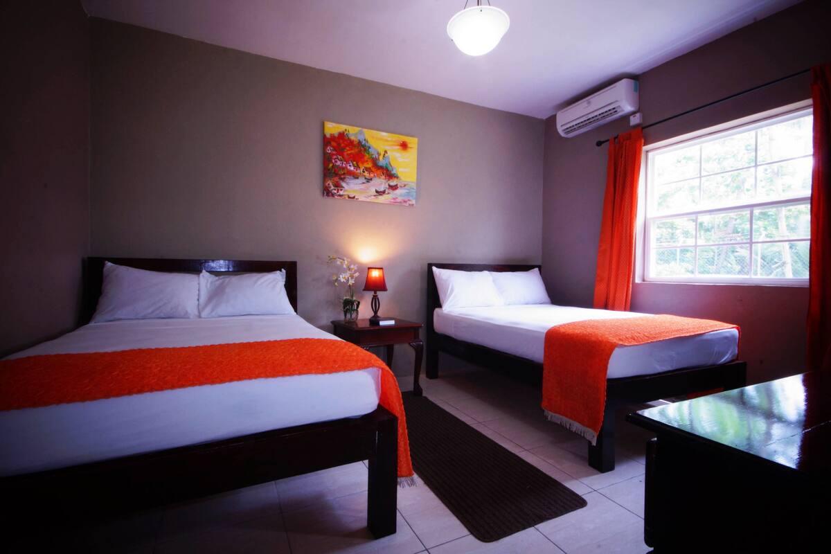 A hotel room with two beds, a nightstand with a lamp, a painting on the wall, and an air conditioning unit.