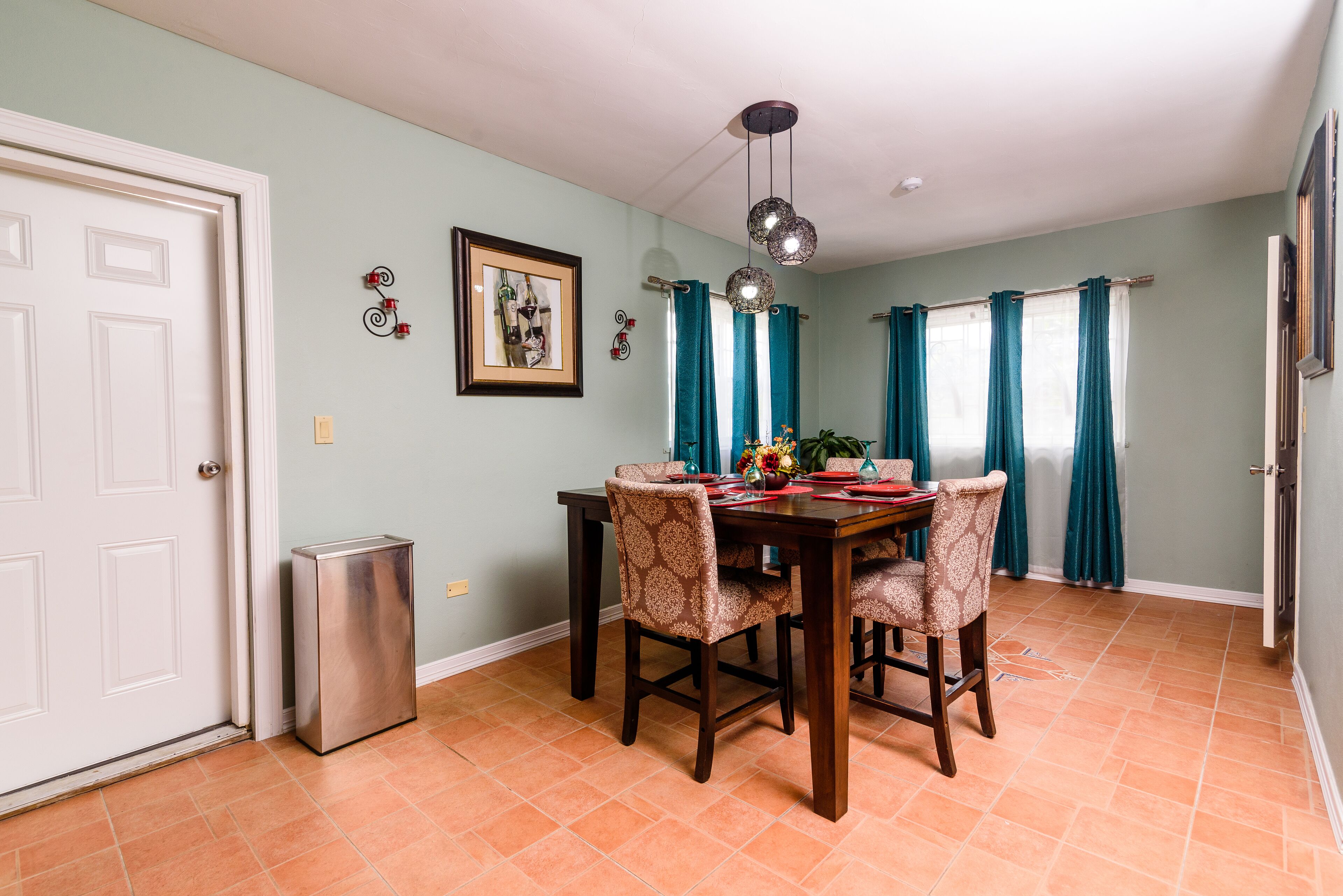 A dining area with a table set for four, a trash can, and teal curtains.